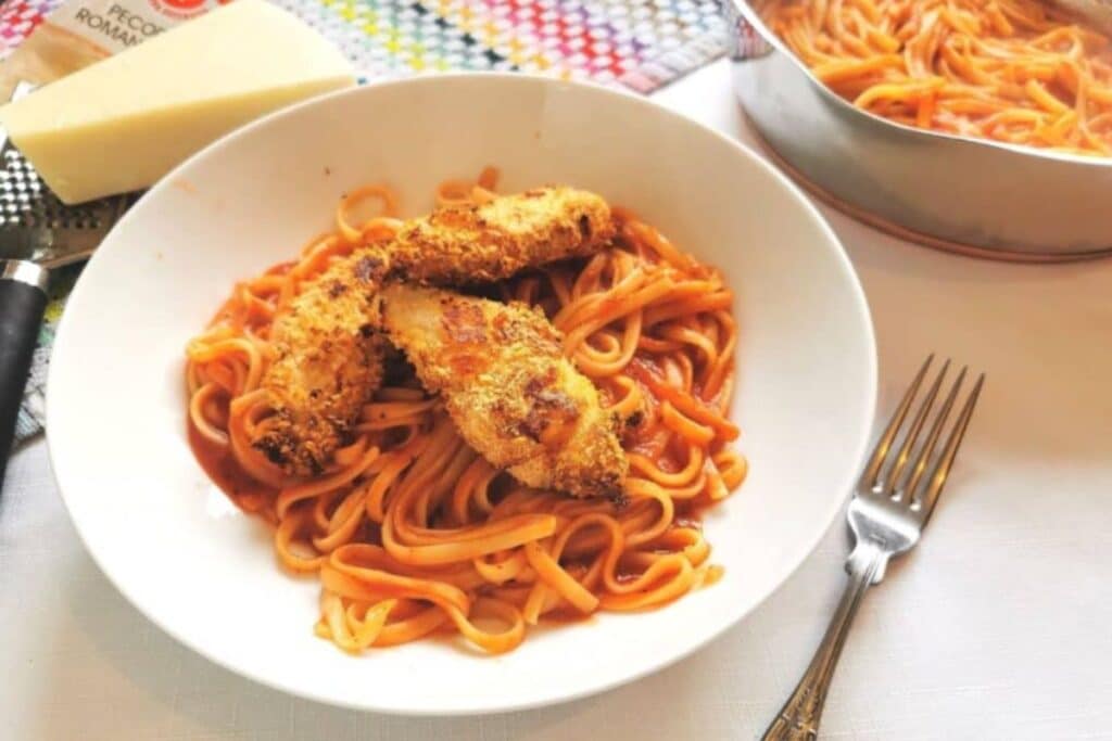 A white bowl with fettuccine pasta in tomato sauce topped with Pecorino Chicken Goujons. A fork is beside the bowl, with a pan of more pasta and a block of cheese in the background.