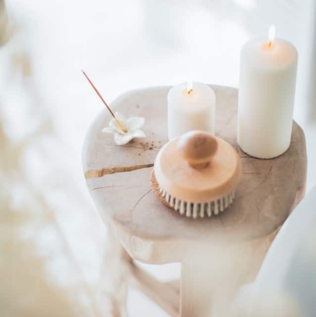 A wooden stool holds two lit white candles, a burning incense stick in a holder, and a wooden dry brush, creating a calm and serene spa-like atmosphere perfect for skincare rituals.