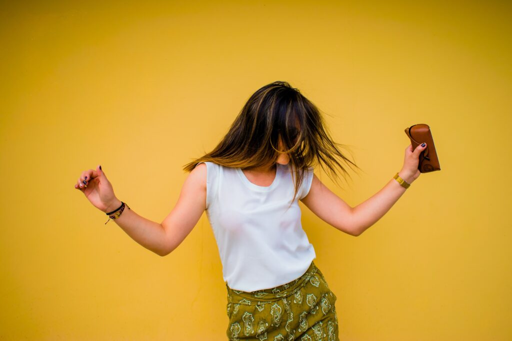 A woman with long hair dances joyfully against a bright yellow wall, wearing a white sleeveless top and green patterned skirt, holding a brown purse—capturing the essence of mood-boosting activities.