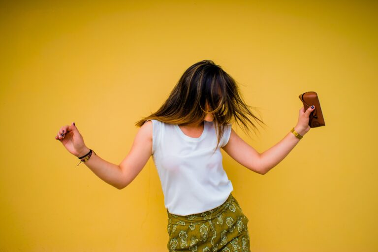 A woman with long hair dances joyfully against a bright yellow wall, wearing a white sleeveless top and green patterned skirt, holding a brown purse—capturing the essence of mood-boosting activities.