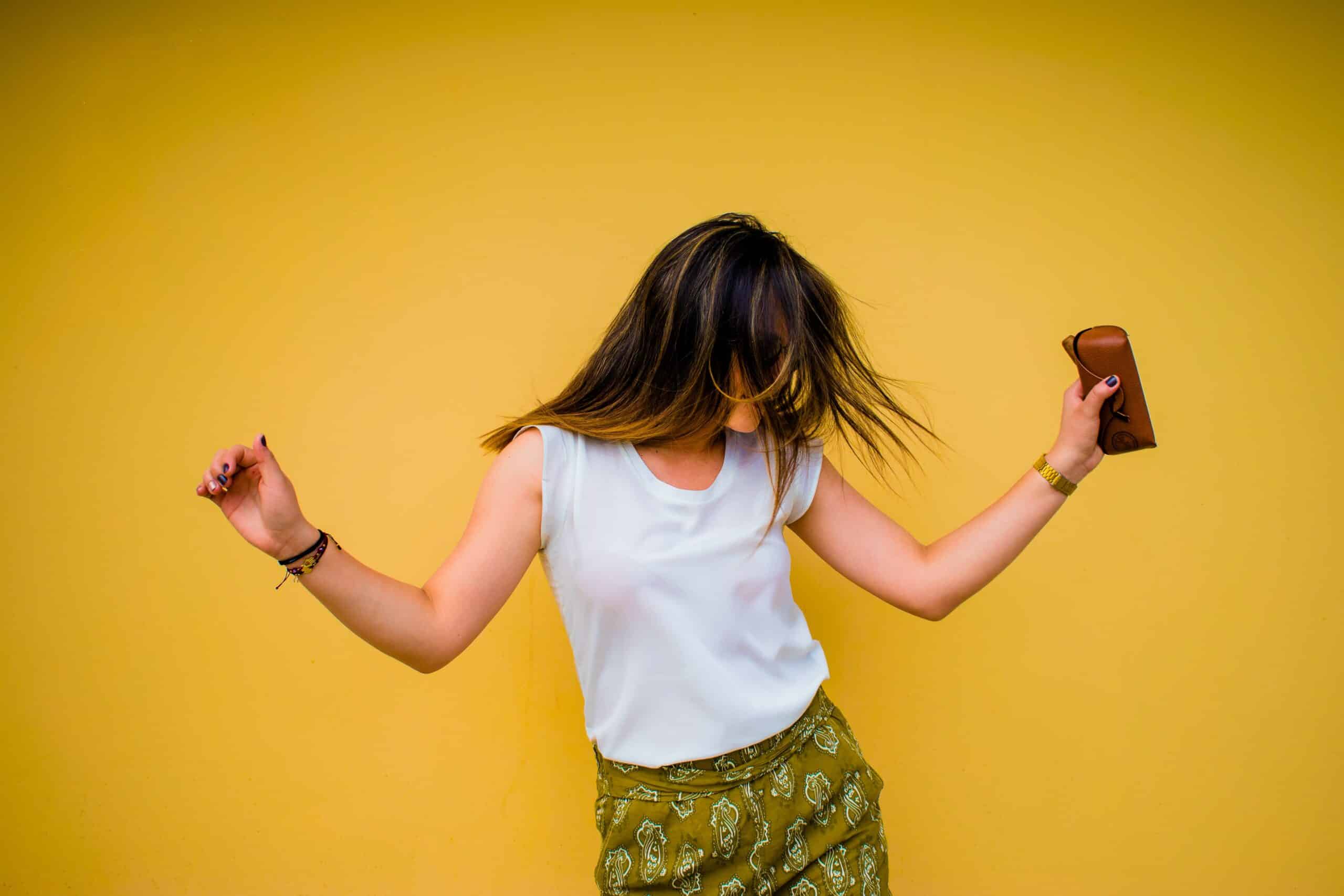 A woman with long hair dances joyfully against a bright yellow wall, wearing a white sleeveless top and green patterned skirt, holding a brown purse—capturing the essence of mood-boosting activities.