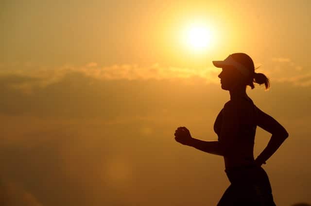 Silhouette of a woman running outdoors at sunrise or sunset, with the sun and clouds in the background. She is wearing a cap and sports attire—showcasing one of many ways to exercise as part of a daily exercise routine.