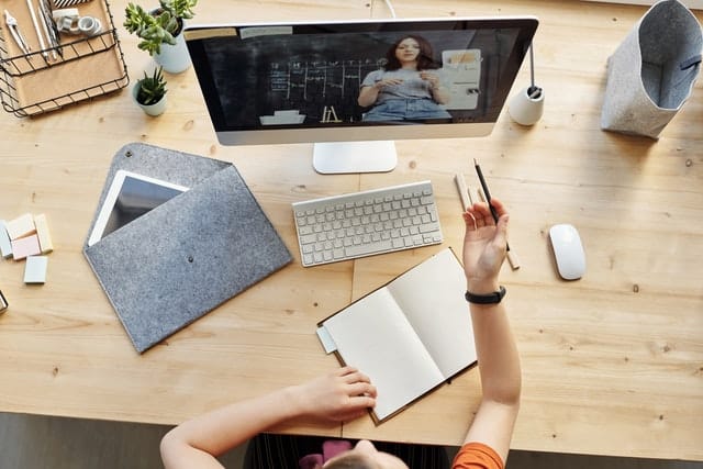 A person sits at a desk with office supplies, a tablet, and a cup, watching a woman give a presentation on the monitor—finding ways to stay connected during the Coronavirus lockdown with notebook, pen, and computer ready on the wooden desk.