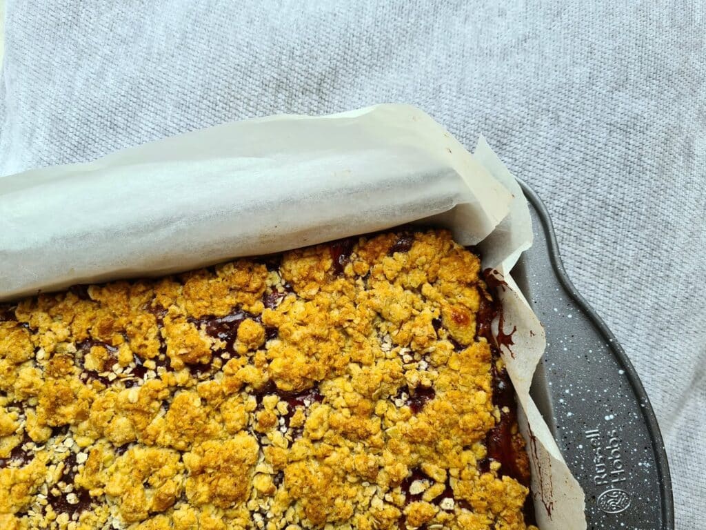 A close-up of a baking tray lined with greaseproof paper, filled with a golden, crumbly oat topping over a fruit pudding, set on a light textured surface—perfect as a photo backdrop for capturing delicious moments.