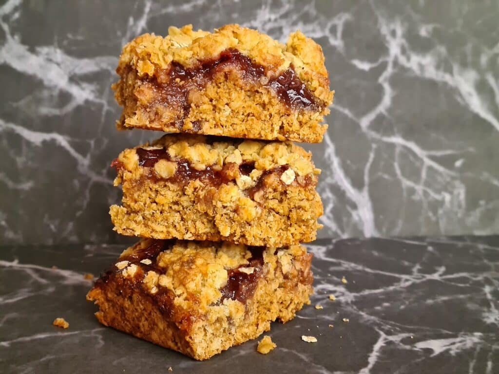 Three stacked oat crumble bars with a layer of dark red jam filling, set against a black and white marble photo backdrop. The bars have a crumbly texture, with scattered crumbs enhancing the visual appeal.