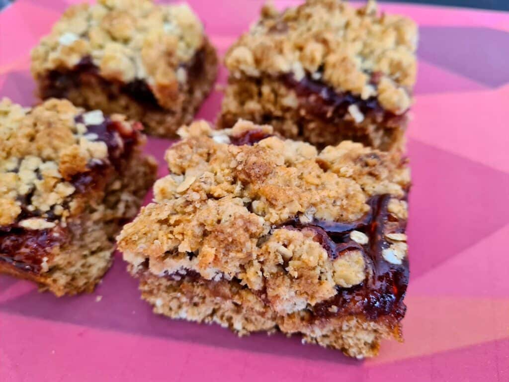 Four oat and jam crumble bars with a golden, crumbly topping and visible layer of red jam, served on a pink geometric-patterned photography background.