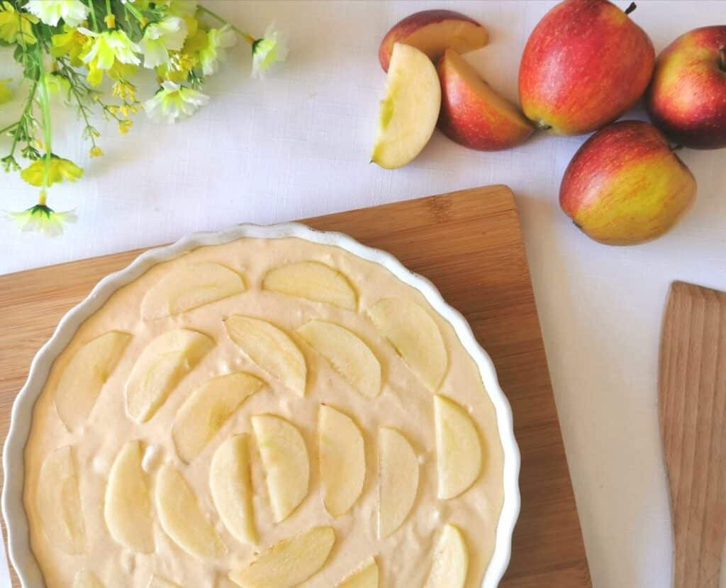 A round baking dish filled with unbaked Jazz Apple Cake mixture, topped with neatly arranged apple slices. Nearby, apples and apple slices rest on a wooden board, with yellow flowers in the corner atop a white tablecloth.