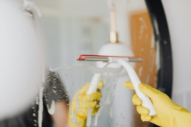A person wearing yellow rubber gloves uses a squeegee to clean a soapy bathroom mirror. A round mirror and a lamp are visible in the background, making this scene perfect for those seeking cleaning tips.
