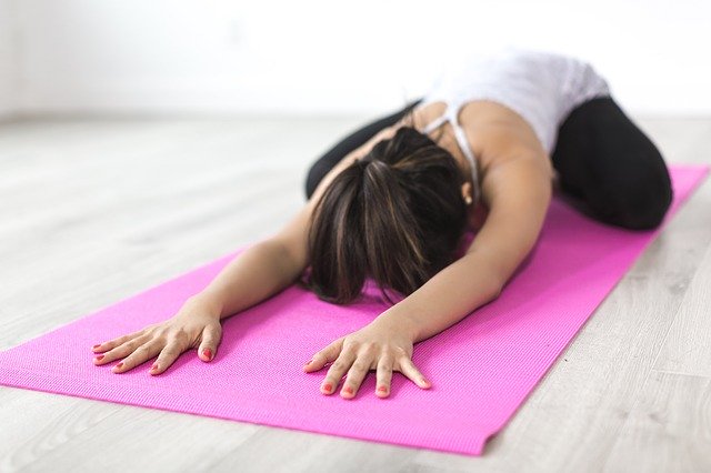 A person in sportswear practises yoga on a pink mat, stretching forward with arms extended and forehead resting on the mat in a bright indoor space, resembling a gentle child’s pose.