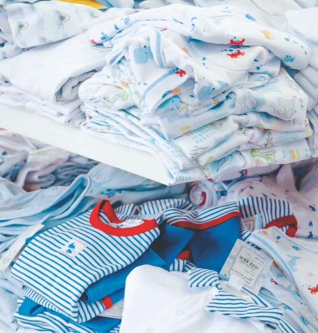 A pile of neatly folded baby clothes, ideal for first-time parents, includes white outfits with colourful prints and blue-and-white striped babygros with red trim, all carefully arranged on shelves.