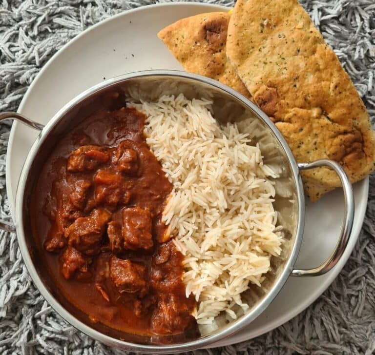 A round metal dish with slow cooker beef curry and white rice, served on a white plate alongside two pieces of naan bread, placed on a textured grey surface.