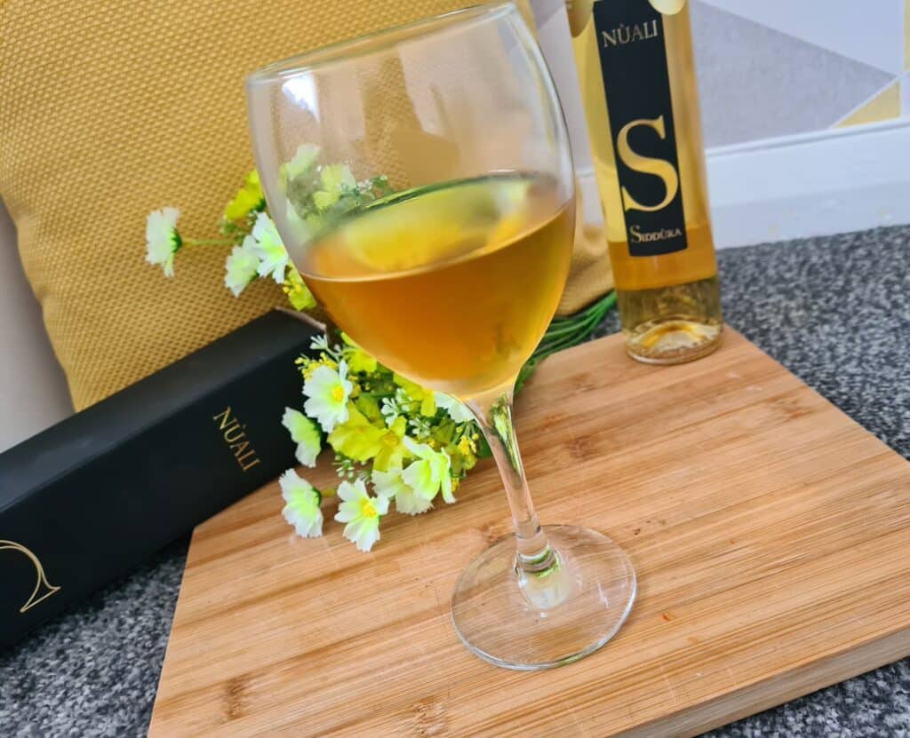 A glass of golden sweet wine sits on a wooden board, paired with a bottle of Nuali S Sweetia Moscato wine, a black box, and yellow-white flowers in the background. The cosy scene invites you to enjoy fruity desserts with a yellow cushion behind.