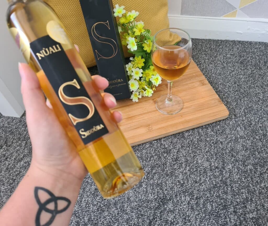 A hand with a triquetra tattoo holds a bottle of Núali S Sundura Moscato wine. In the background, there's a glass of the same wine on a wooden tray with yellow flowers and the wine's black and gold box—perfect for fruity desserts and wine pairing.