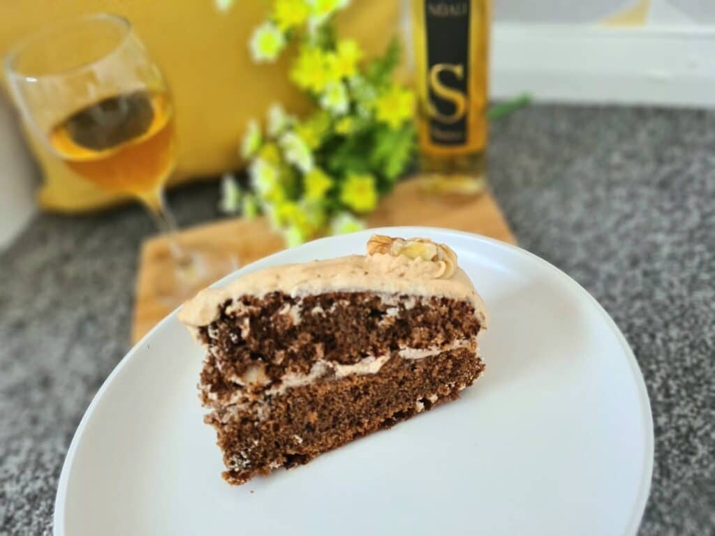 A slice of frosted coffee and walnut cake sits on a white plate. In the background, a glass of Moscato wine adds a touch of sweetness—perfect for those who enjoy unique wine pairings with fruity puddings and classic cakes alike.