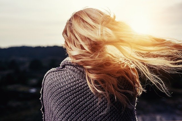 A person with long blonde hair, wearing a knitted jumper, stands outdoors with their back to the camera. Their hair is blowing in the wind as the sun sets—an ideal scene to remember key Hair Protection Tips for glowing summer hair.