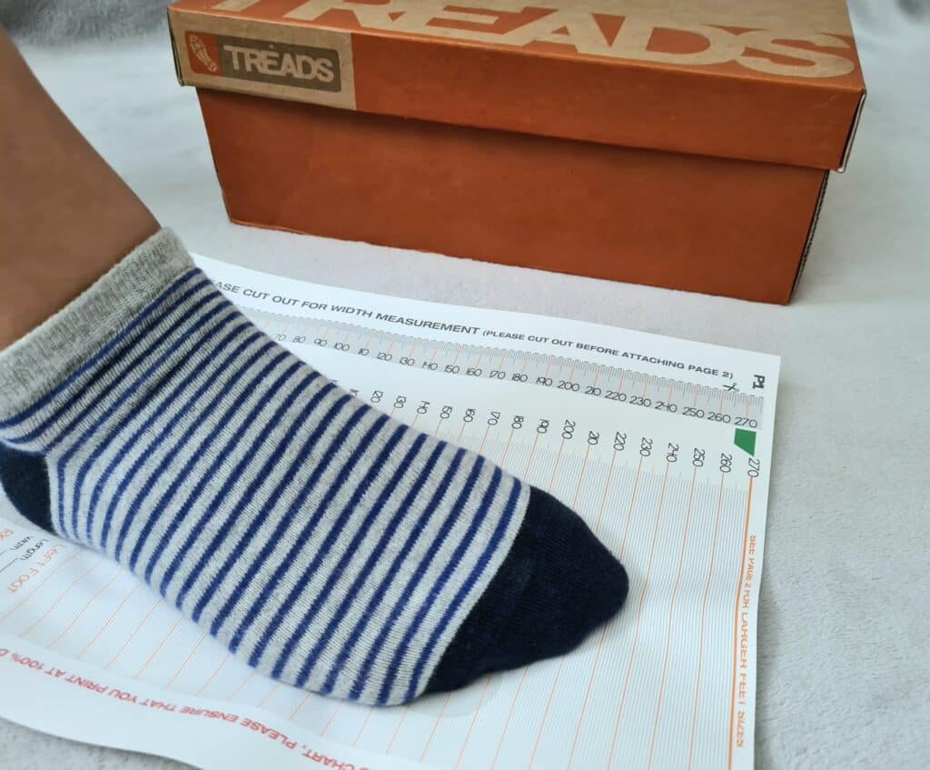 A foot wearing a blue and white striped sock is placed on a shoe size measurement chart, ready for fitting school shoes. In the background, an orange shoe box labelled "TREADS" is visible.