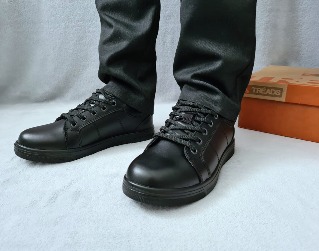 A person wearing black lace-up school shoes and dark trousers stands on a light grey surface, with an orange shoe box labelled "Treads" in the background.