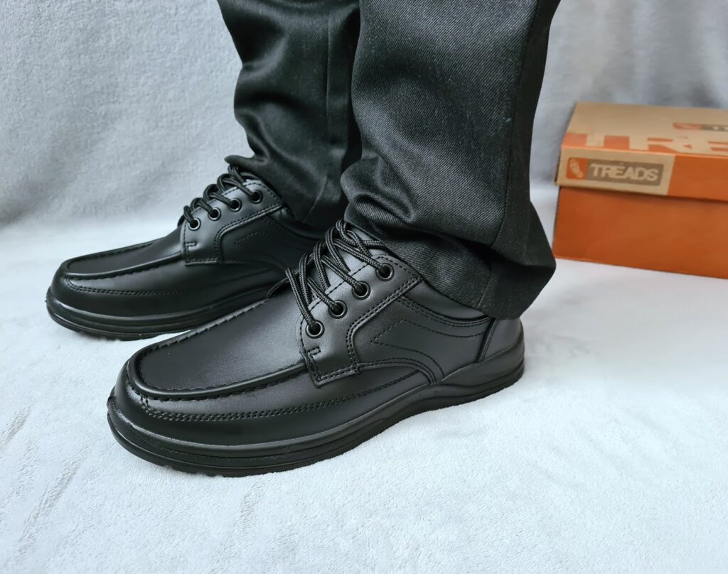 A person wearing black leather school shoes and black trousers stands on a light surface; an orange shoe box labelled "TREADS" is visible in the background.