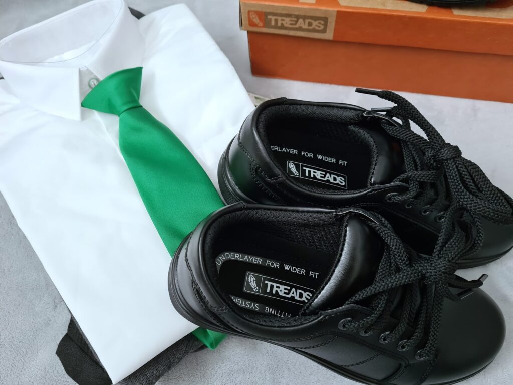 A white shirt with a bright green tie is neatly folded next to a pair of black lace-up school shoes. Behind them are two stacked orange and brown boxes labelled "Treads.