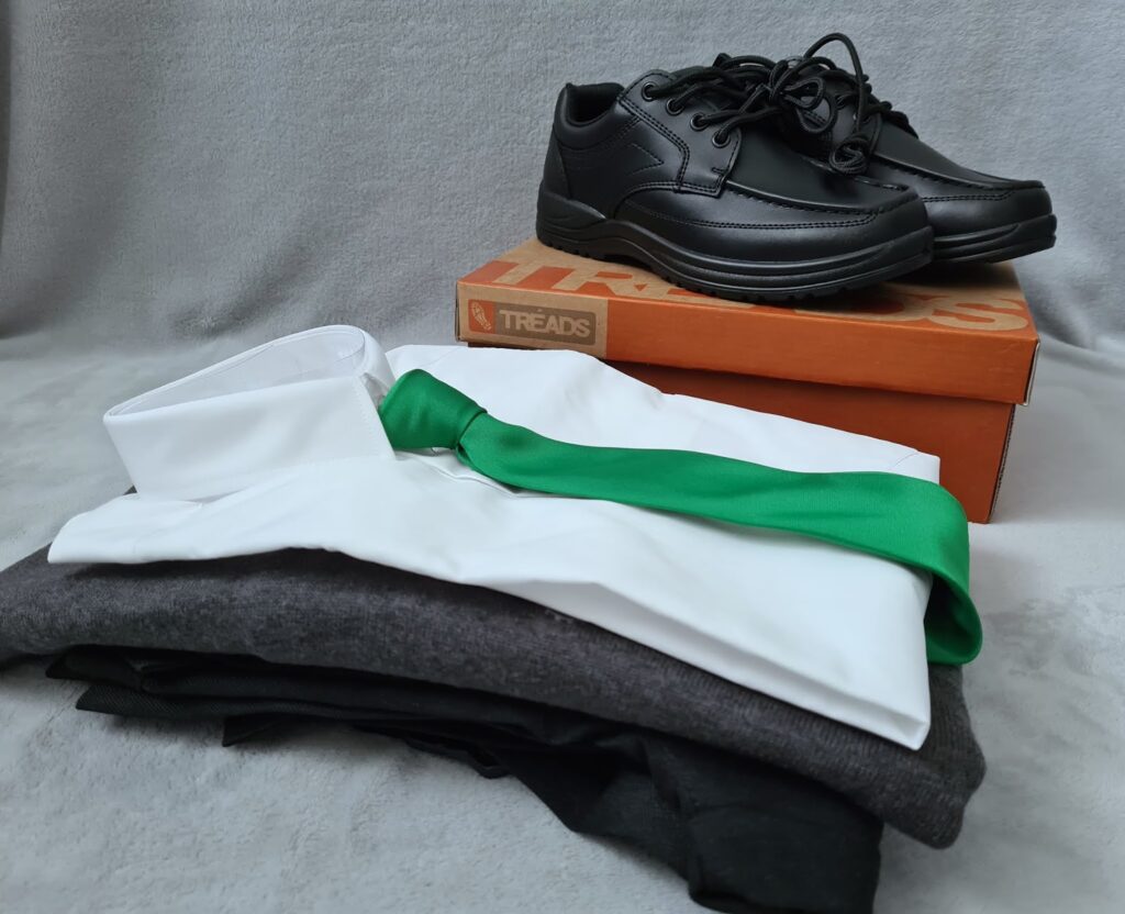 A pair of black school shoes sits on an orange shoebox behind a neatly folded white dress shirt with a green tie, stacked atop a grey jumper and black trousers, all arranged on a light grey surface.