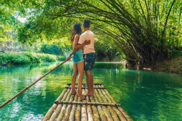 A couple stands on a bamboo raft floating down a lush, green river, surrounded by overhanging trees and sunlight filtering through the leaves—one of many picturesque family holiday destinations.