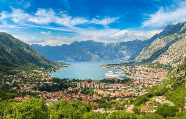A panoramic view of a coastal town surrounded by mountains, with red-roofed buildings, lush greenery, and a cruise ship docked in the bay—an idyllic scene perfect for top family holiday destinations under a bright blue sky with scattered clouds.