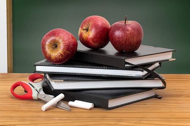 Three red apples rest on a stack of black-covered books, with red-handled scissors and two pieces of white chalk beside them, all set on a wooden desk in front of a green blackboard, highlighting the importance of pastoral care in schools.