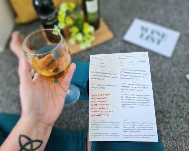 A person holding a glass of white wine sits on the floor with a wine-tasting menu in hand, surrounded by my favourite things—two bottles, flowers, and a card reading "WINE NOT?" A tattoo adorns their wrist.