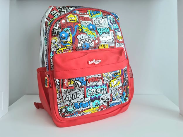 A colourful backpack with comic-style designs and words like "POW!", "ZAP", and "SPLASH" on the front. The main body is red, with a large front pocket displaying the Smiggle brand logo—perfect for carrying all my favourite things.