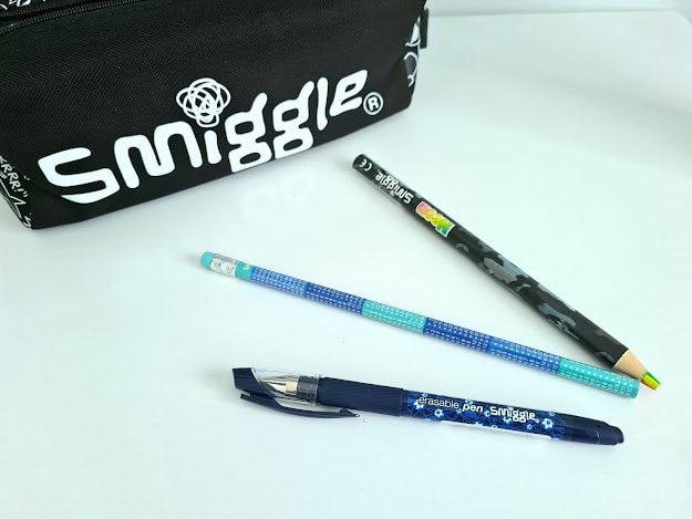 A black Smiggle pencil case sits next to a blue pen, a blue patterned pencil, and a camouflage-patterned pencil—some of my favourite things—on a white surface.