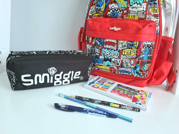 A colourful comic-themed Smiggle backpack stands behind a matching black Smiggle pencil case. In front are two pencils, a pen, and a colouring book—perfect for keeping all my favourite things organised on a white surface.