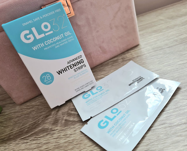 A box of Glo32 Advanced Whitening Strips with coconut oil sits on a wooden surface next to two opened sachets and a pink pouch—one of my favourite things for enamel-safe, effective whitening. The packaging highlights its benefits.