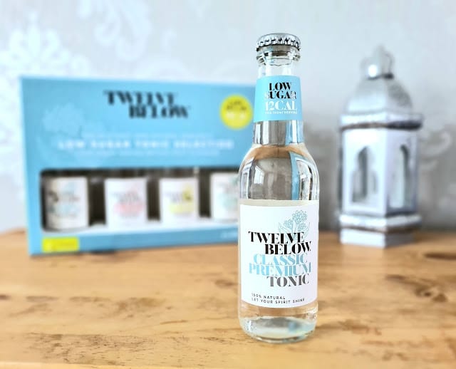 A clear bottle of TwelveBelow Classic Premium Tonic, known for its low-sugar tonics, sits on a wooden surface. Behind it, a blue box with more tonic bottles and a white lantern add a softly blurred background.