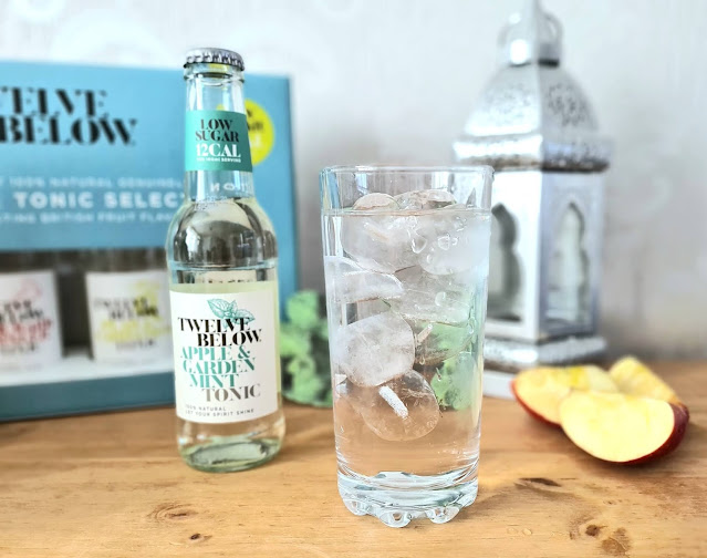 A glass filled with ice and clear low sugar tonic water sits on a wooden surface beside a bottle of "Twelve Below Apple & Garden Mint Tonic." Nearby are apple slices and a product box in the background.