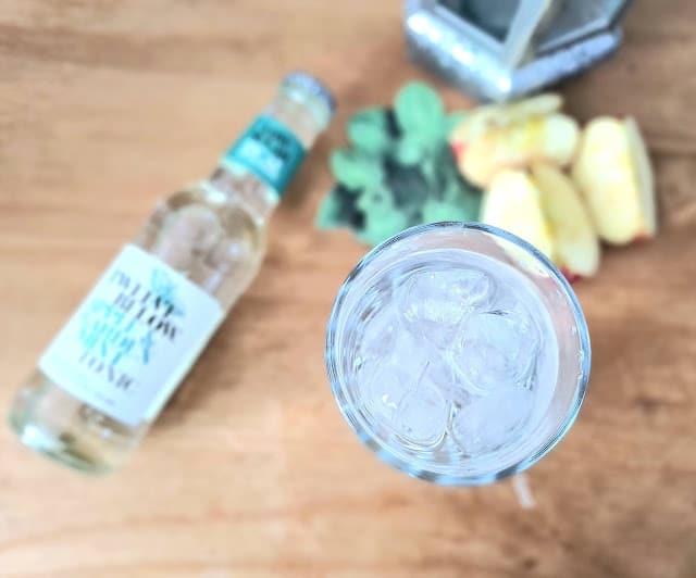 A glass filled with ice cubes sits on a wooden surface next to a bottle of TwelveBelow low calorie tonics, with mint leaves and sliced apples slightly out of focus in the background.