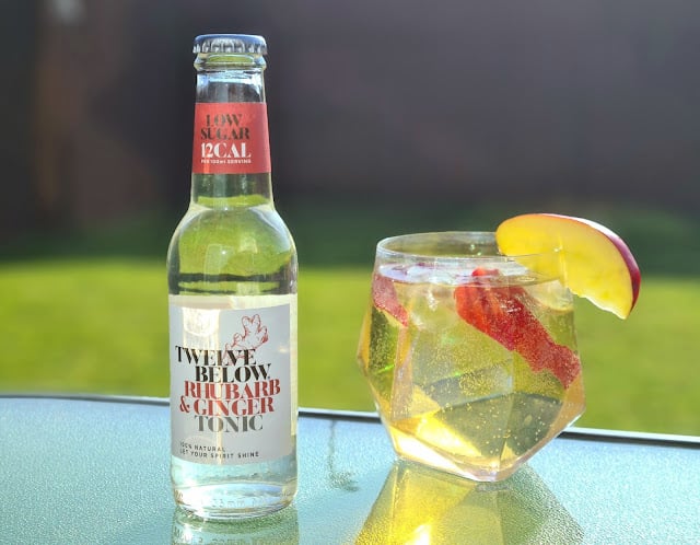 A bottle of TwelveBelow Rhubarb & Ginger Tonic, known for its low sugar tonics, stands beside a faceted glass with tonic, ice, red apple, and strawberry slices on a glass table with green grass in the background.