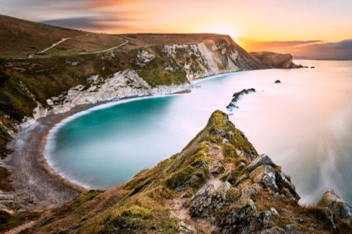 A curved bay with turquoise water and steep, grassy cliffs at sunset—an idyllic spot for a family travel adventure. Warm orange hues light the sky as rocky formations stretch into the calm sea along the shoreline.