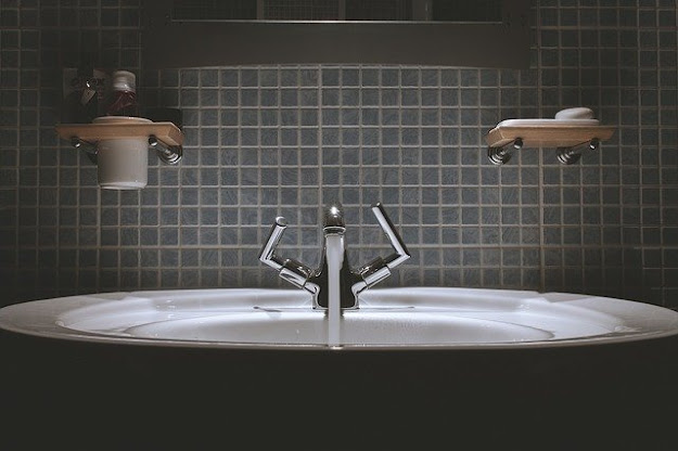 A close-up view of a bathroom sink with a modern chrome tap, set against dark mosaic tiles—perfect inspiration for Bathroom Remodelling Ideas. Shelves above the sink hold a mug, toiletries, and a soap dish.