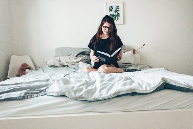 A woman with long hair and glasses sits cross-legged on an unmade bed in a bright, minimal bedroom, reading about mattress durability. A small framed leaf print hangs on the wall above her.
