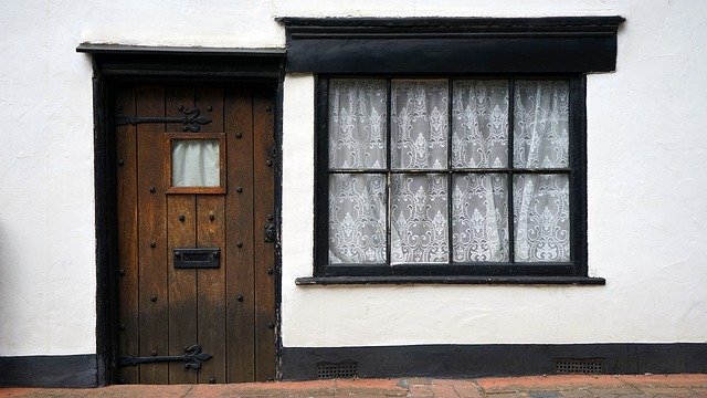 A wooden front door with a small window sits beside a large, lace-curtained window on a white plastered house, framed in black—creating charming kerb appeal sure to attract potential buyers as the pavement stretches below.