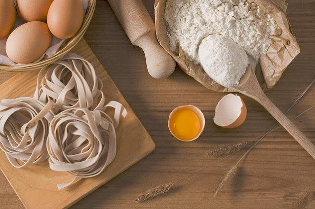 A wooden table with raw pasta nests, whole eggs in a basket, flour in a paper bag with a wooden spoon, a rolling pin, and a cracked egg showing the yolk—has the pandemic made the UK fall back in love with tradition?.