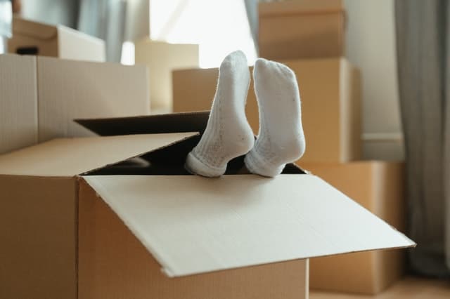 A person wearing white socks has their legs sticking out of an open cardboard box, surrounded by other moving boxes in a room with soft natural light—a candid snapshot of moving house.