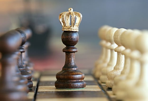 A dark wooden chess pawn wears a gold crown and stands between two rows of chess pieces on a chessboard, suggesting the concept of potential or transformation—much like earning an Amazon Voucher through skilful play.