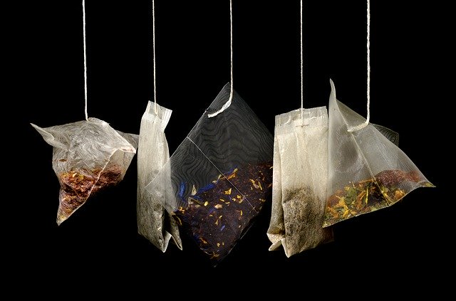 Four tea bags filled with different types of loose teas hang by strings against a black background. Each bag offers a unique blend, perfect for morning tea or night tea, showcasing an array of colours and textures.