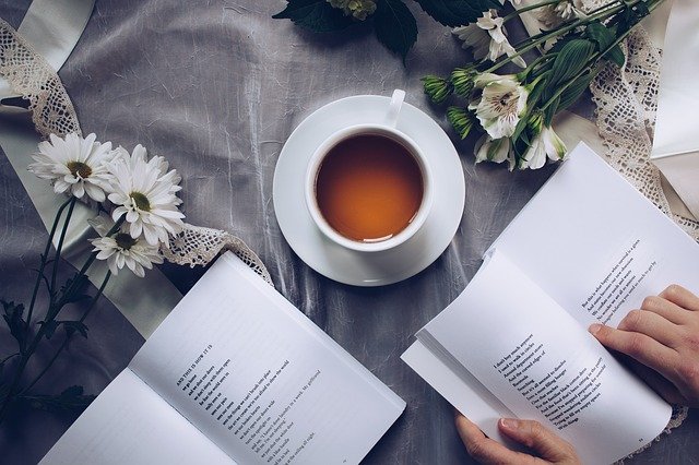 A cup of morning tea sits on a table next to white flowers, lace, and two open books, whilst a person’s hands flick through one of the books. The scene feels calm and cosy.