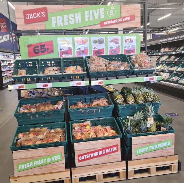 A supermarket display labelled "Fresh Five" showcases crates of fresh fruit and vegetables, perfect for budget friendly shopping. Signs read "Outstanding Value," "Every Fortnight," with price labels visible on pineapples, potatoes, onions, and apples.