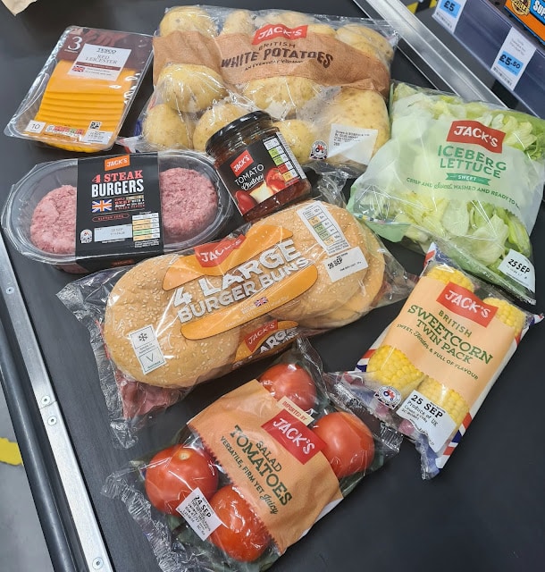 A selection of grocery items on a checkout belt, including potatoes, beef burgers, cheese slices, burger buns, tomatoes, sweetcorn, iceberg lettuce, and tomato relish—mostly from Jack's brand—for budget-friendly shopping.