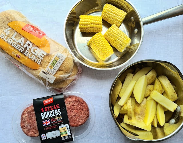 A pack of four burger buns, four raw beef burgers, sliced potatoes in a saucepan, and corn on the cob pieces—all arranged on a white surface—perfect for budget-friendly shopping and easy family meals.
