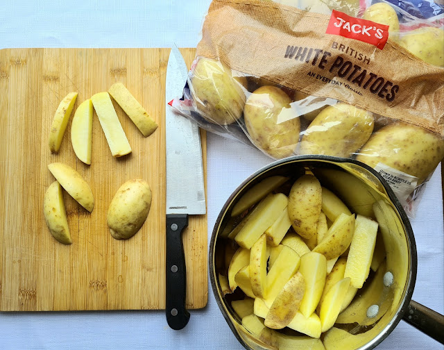 A chopping board with sliced potatoes and a knife, a saucepan filled with potato wedges, and a bag of Jack’s British White Potatoes on a white surface—perfect for budget-friendly shopping.