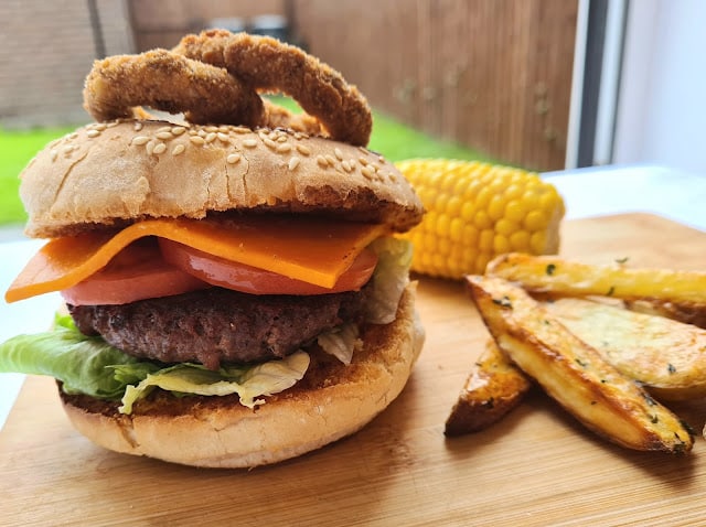 A budget-friendly shopping favourite: a cheeseburger with lettuce, tomato, cheddar cheese, and onion rings in a sesame seed bun, served with potato wedges and corn on the cob on a wooden board.